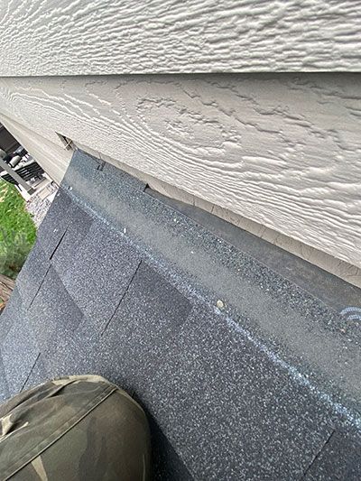 Close-up of roof shingles meeting a white siding wall, with a shoe visible at the bottom left.