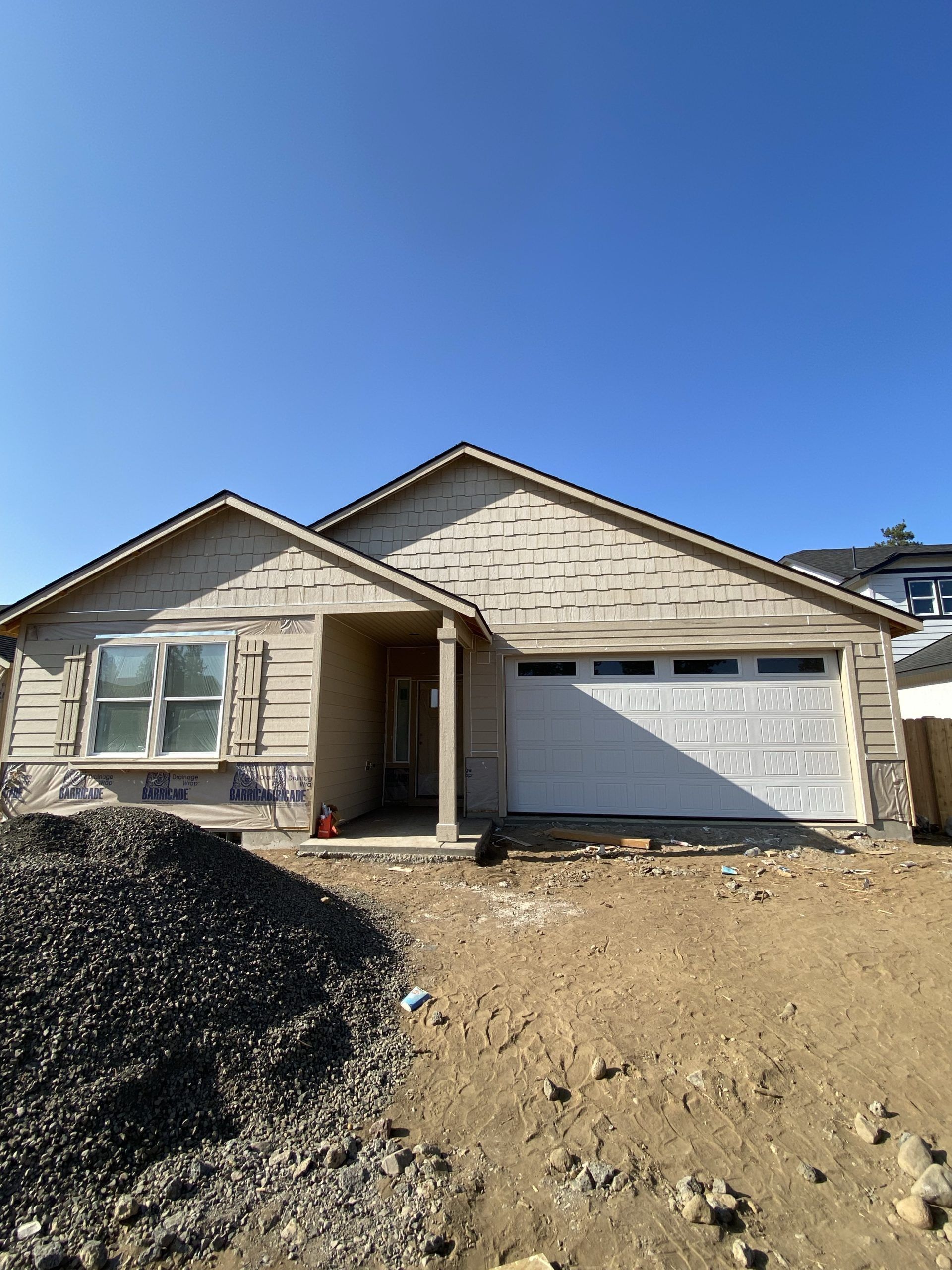 New house under construction with stone facade, garage, and dirt front yard under a clear blue sky