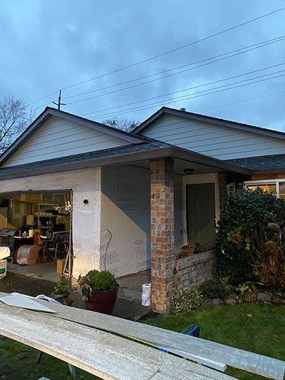 Single-story house with open garage, front porch, and stacked lumber in the yard under a cloudy sky