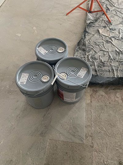 Three gray buckets with lids on a concrete floor near a tarp and red stand.