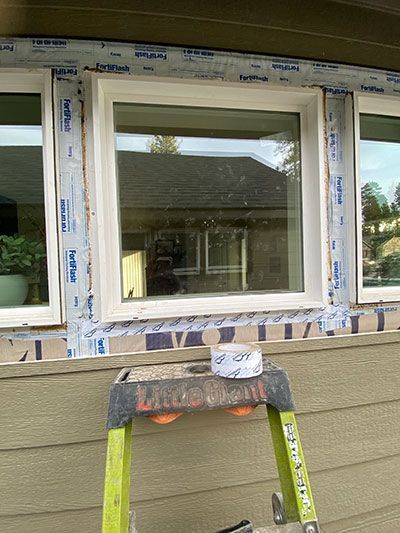Window being installed on house exterior, with ladder and siding wrapped in protective tape.
