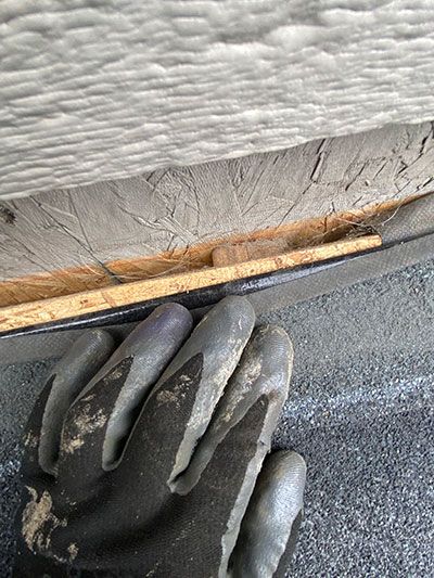 Gloved hand scraping old caulk along a wall seam near trim and insulation.