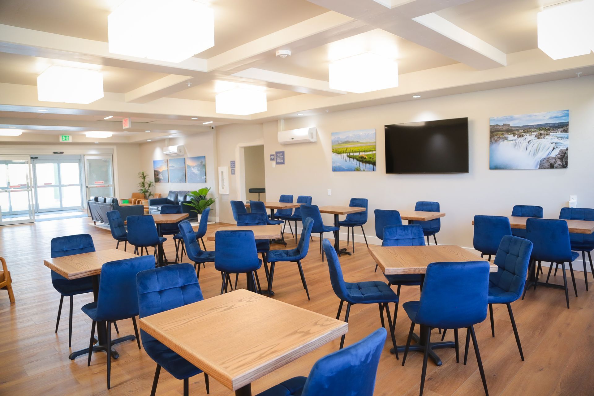 Bright cafeteria with blue chairs, wooden tables, ceiling lights, wall art, and a TV screen.
