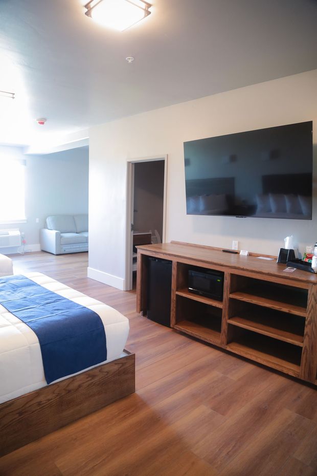 Modern bedroom with a blue bed, wall-mounted TV, wood floor, and a small sitting area in the background
