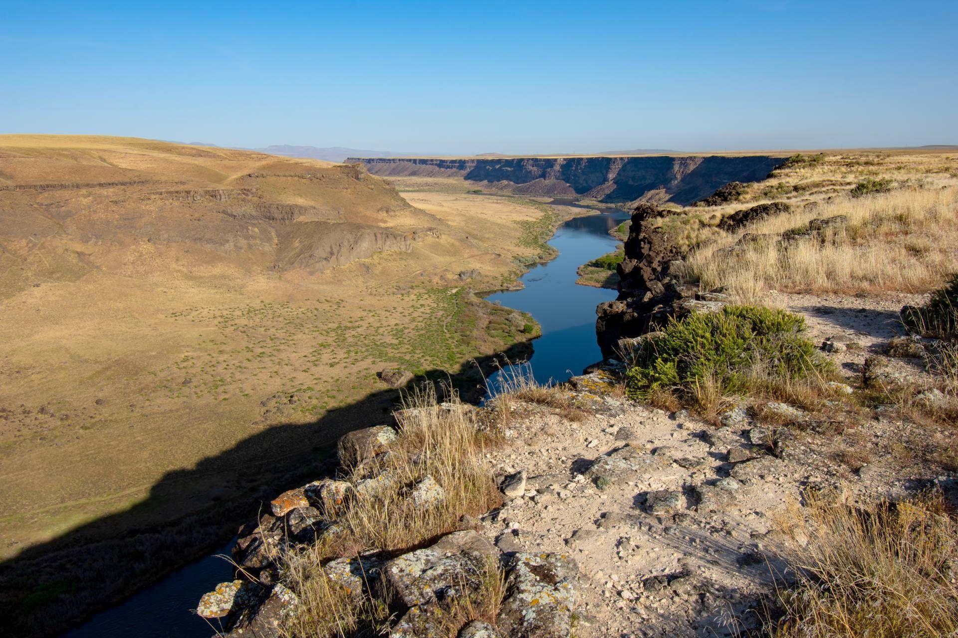 Canyon landscape with a river winding through it. Brown cliffs, blue water, and dry vegetation under a clear sky.