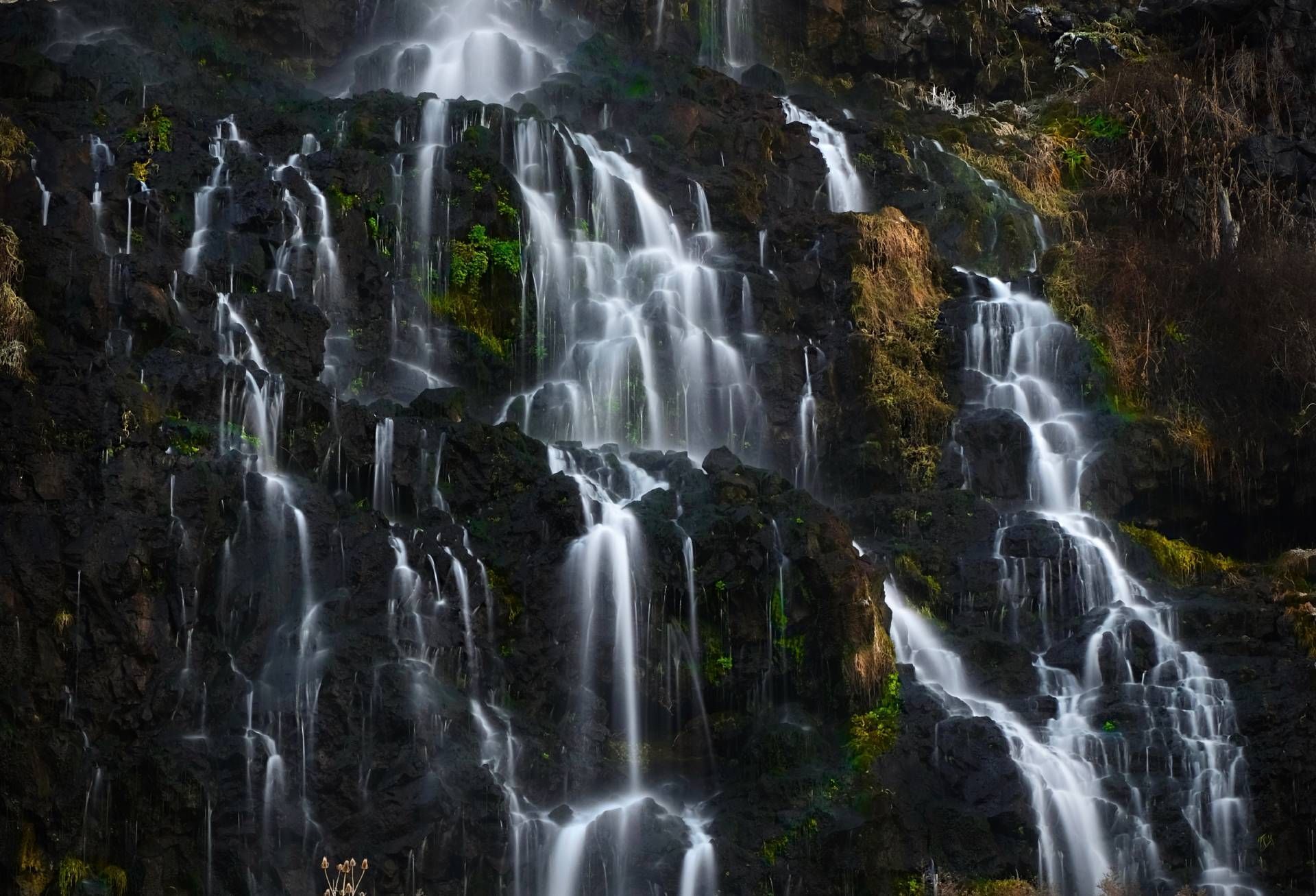 Multiple waterfalls cascading down a dark, rocky hillside; white water against dark stone.