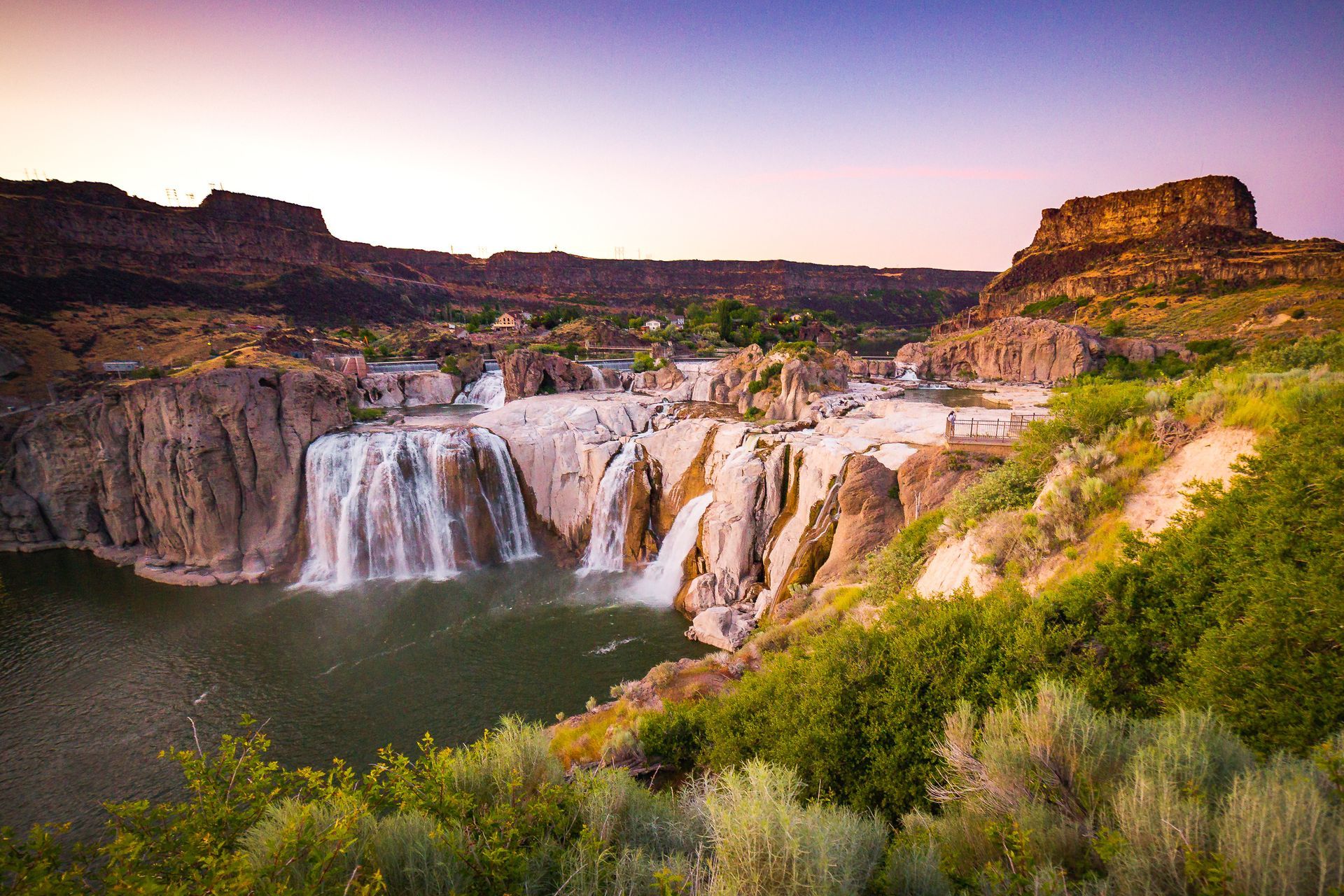 Waterfall cascading into a river canyon, framed by rock formations and greenery, under a colorful sunset.