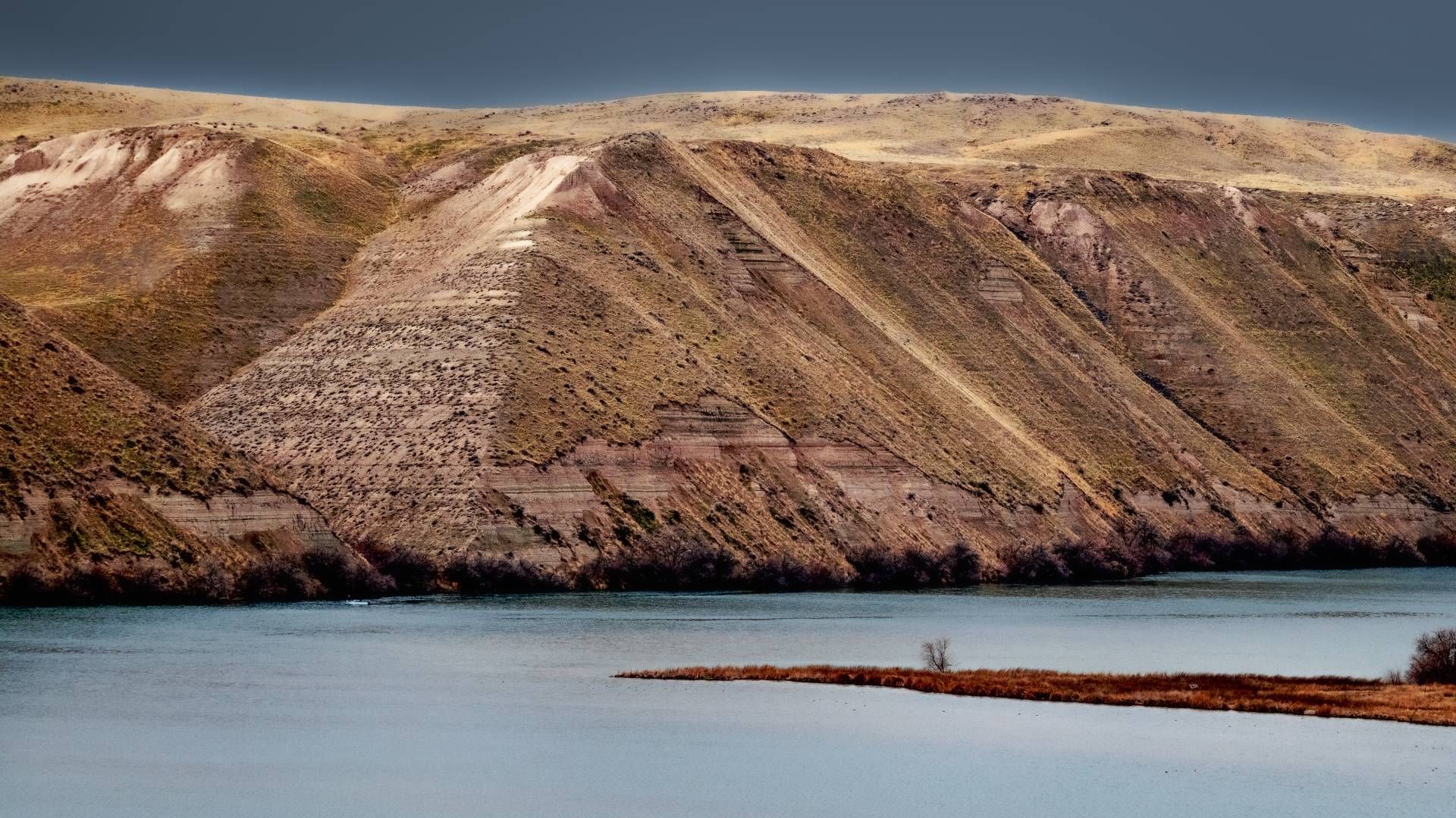 Brown, terraced hills rise above blue water, under a cloudy sky.