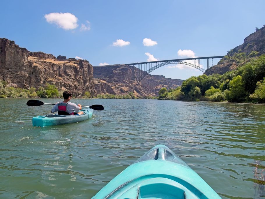 Kayakers on river under bridge in canyon, blue sky.