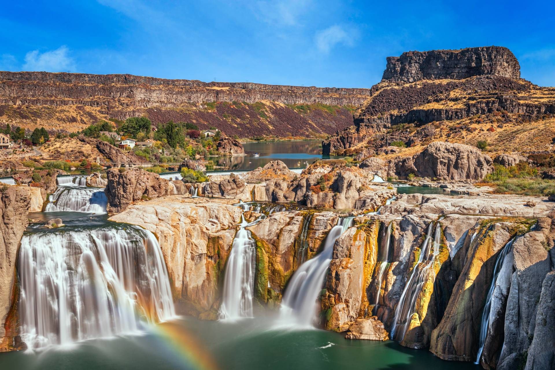 Waterfall with rainbow over turquoise water, brown cliffs in background, and blue sky.