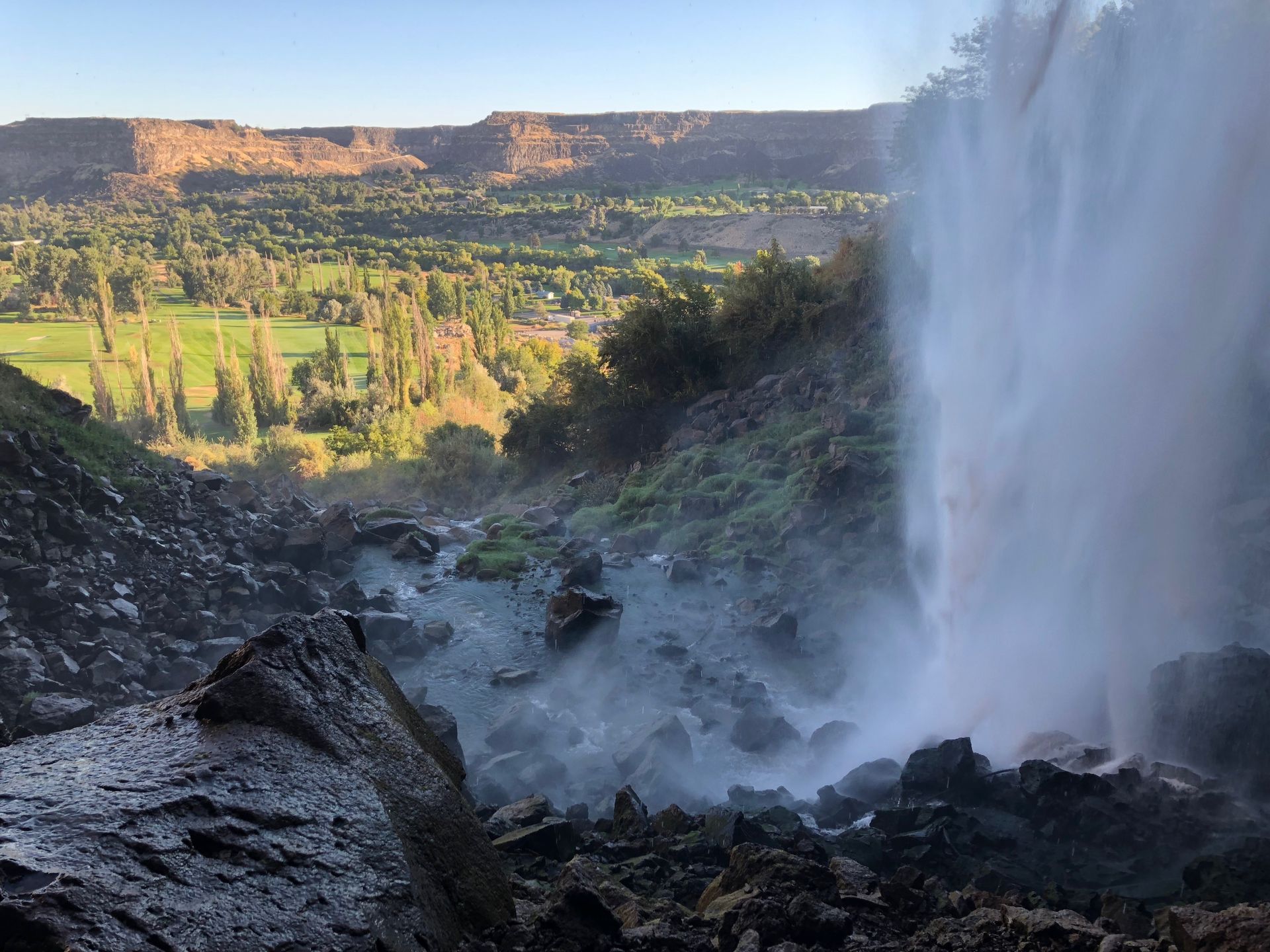 Waterfall cascading over dark rocks into a misty canyon with green valley and cliffs beyond.