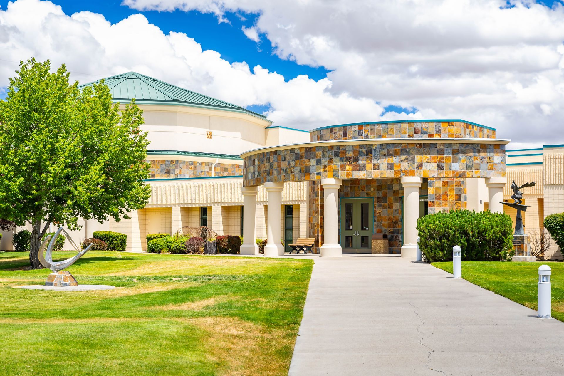 Modern building entrance with columns, curved stone facade, and a walkway amid green lawn and trees