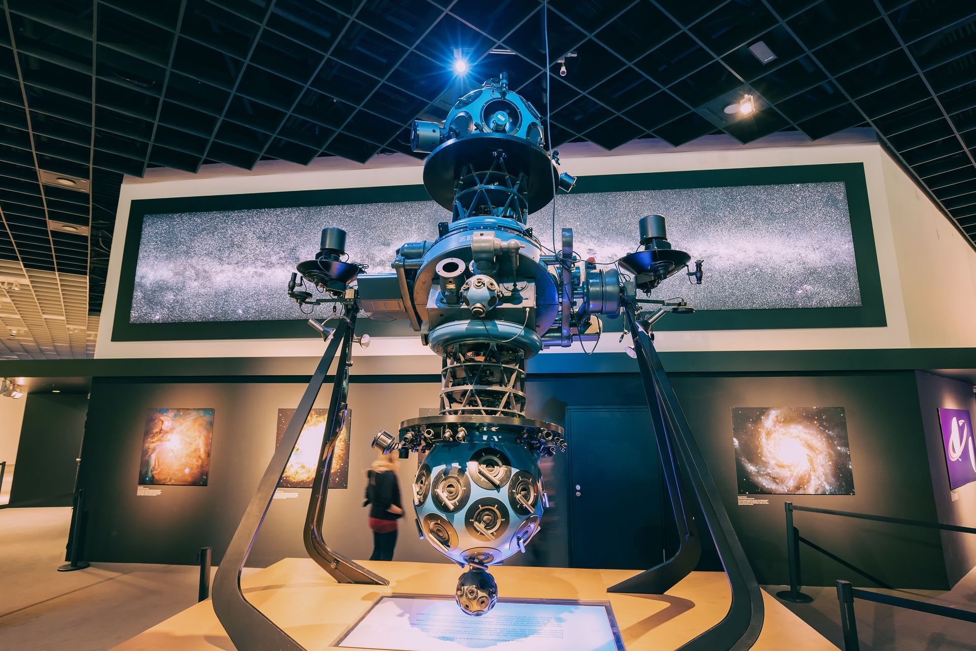 Large blue robotic sculpture on display in a museum gallery with spotlights and framed wall art