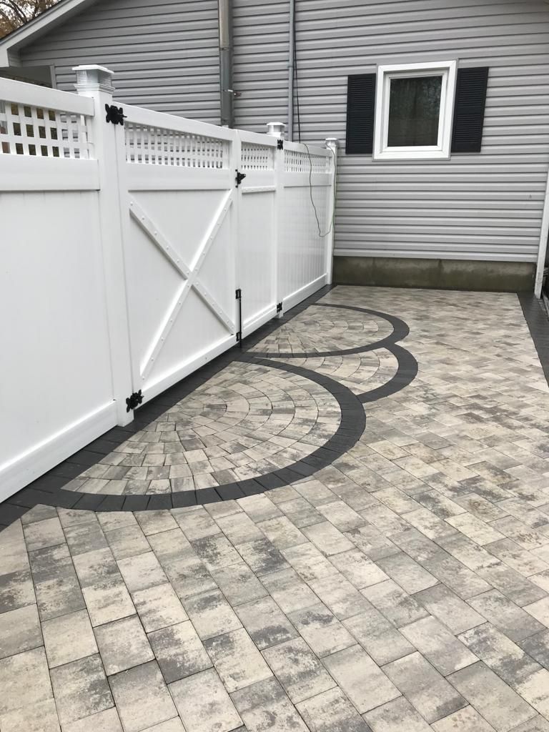 White vinyl fence and gate beside a gray house, with a patterned paver driveway and curved dark inlay.
