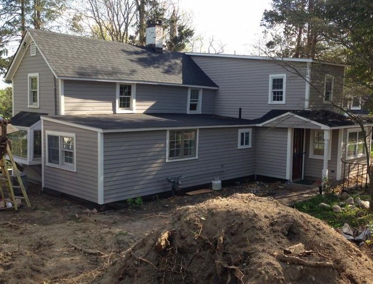 Gray two-story house with new siding, surrounded by bare dirt and trees on a sloped lot
