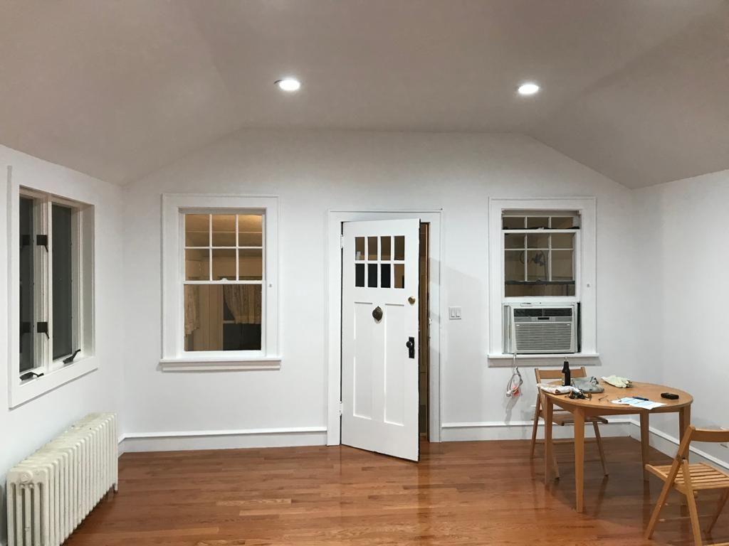 Bright empty room with white walls, wood floor, windows, a door, and small table with chairs.