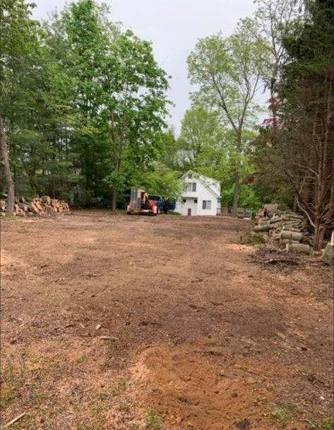 Cleared dirt driveway leading to a small white house, with trees and a construction machine nearby