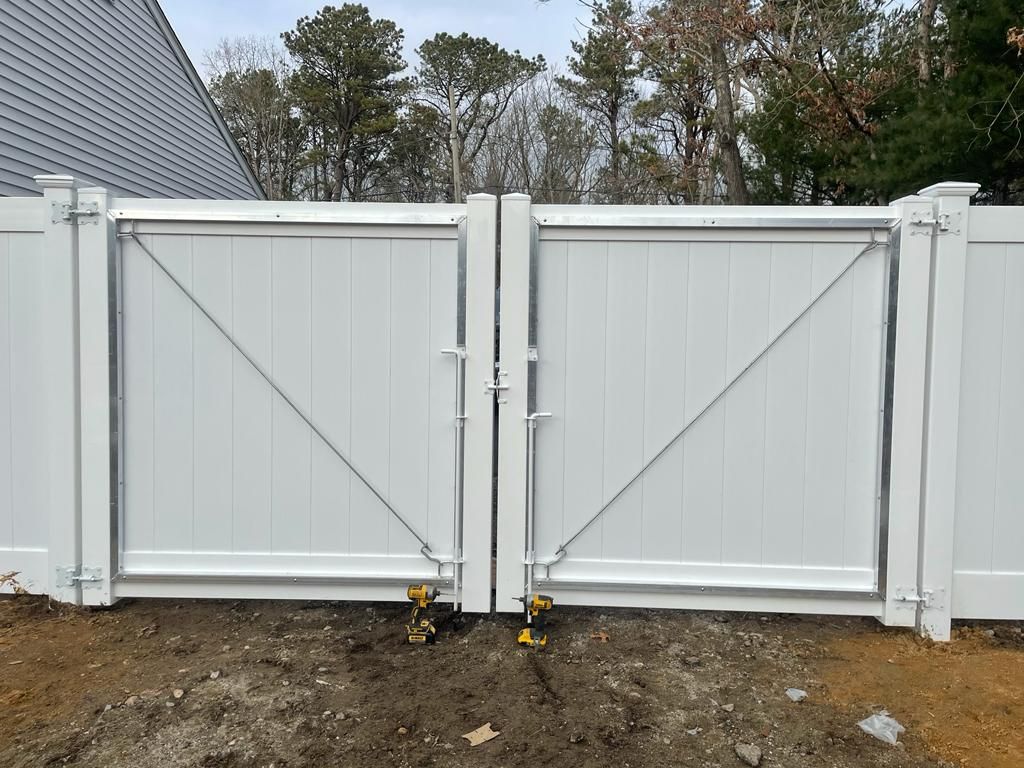Closed white double gate with diagonal braces in a fenced yard, with dirt ground and trees behind it