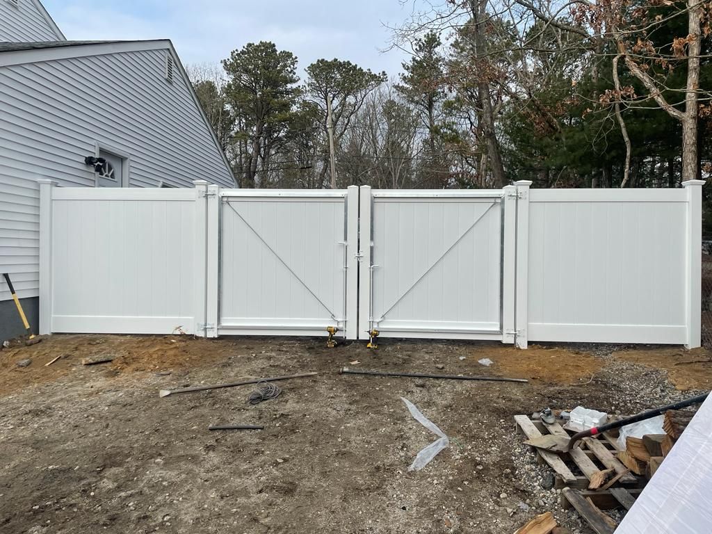 White wooden double gate in a fenced yard beside a house, with trees and dirt ground in front.