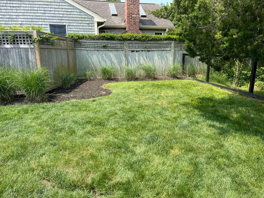 Backyard lawn with wooden fence, shrubs, and trees behind a house