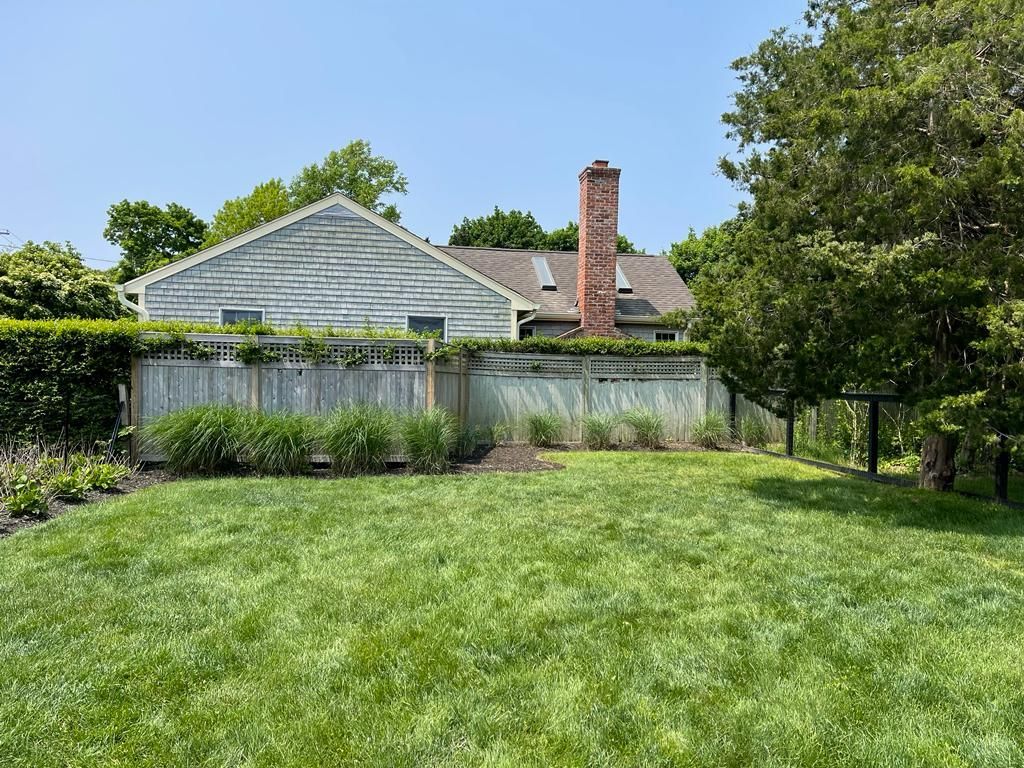 Backyard with green lawn, trees, and a house with a chimney under a clear blue sky