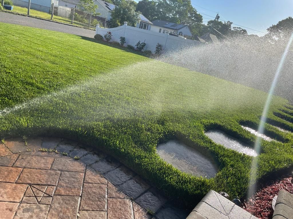 Sprinklers watering a green lawn beside a brick patio in bright sunlight