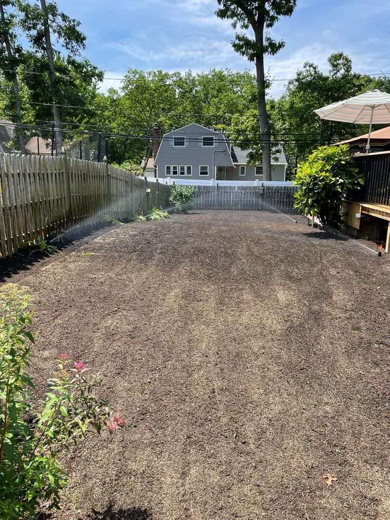Gravel driveway or yard bordered by fences and trees, with a house in the background under a bright sky
