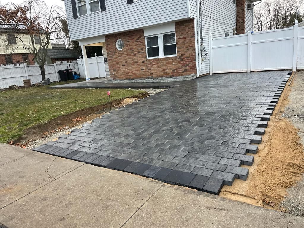 Freshly paved dark-gray driveway beside a house with a brick wall and white fence.