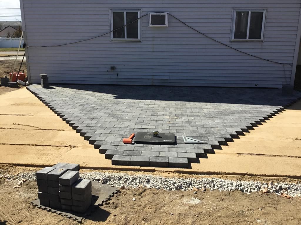 Partially paved patio with gray pavers in front of a house, with stacked bricks and tools nearby