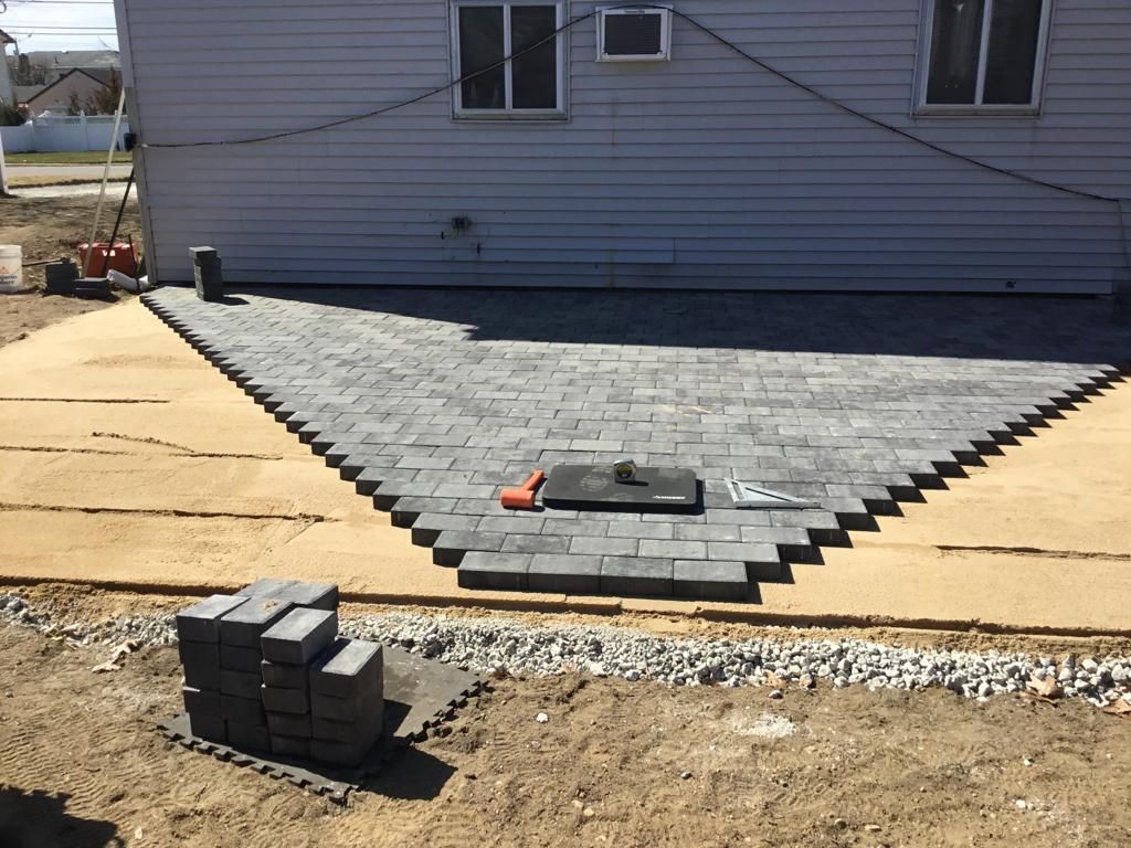 Partially paved front yard with a gray brick walkway, tan pavers, and a stack of bricks beside a house.