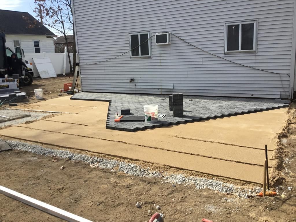 Backyard patio under construction beside a gray house, with concrete forms and gravel in the foreground