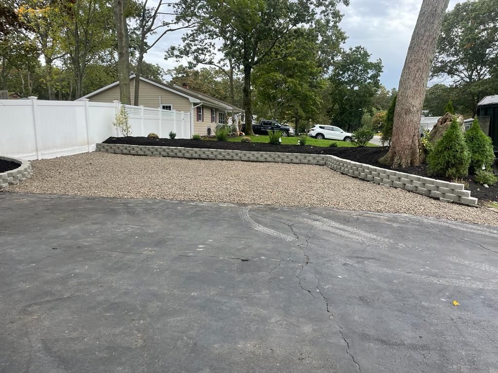 Gravel driveway beside a house, with a white fence, trees, and a paved foreground.