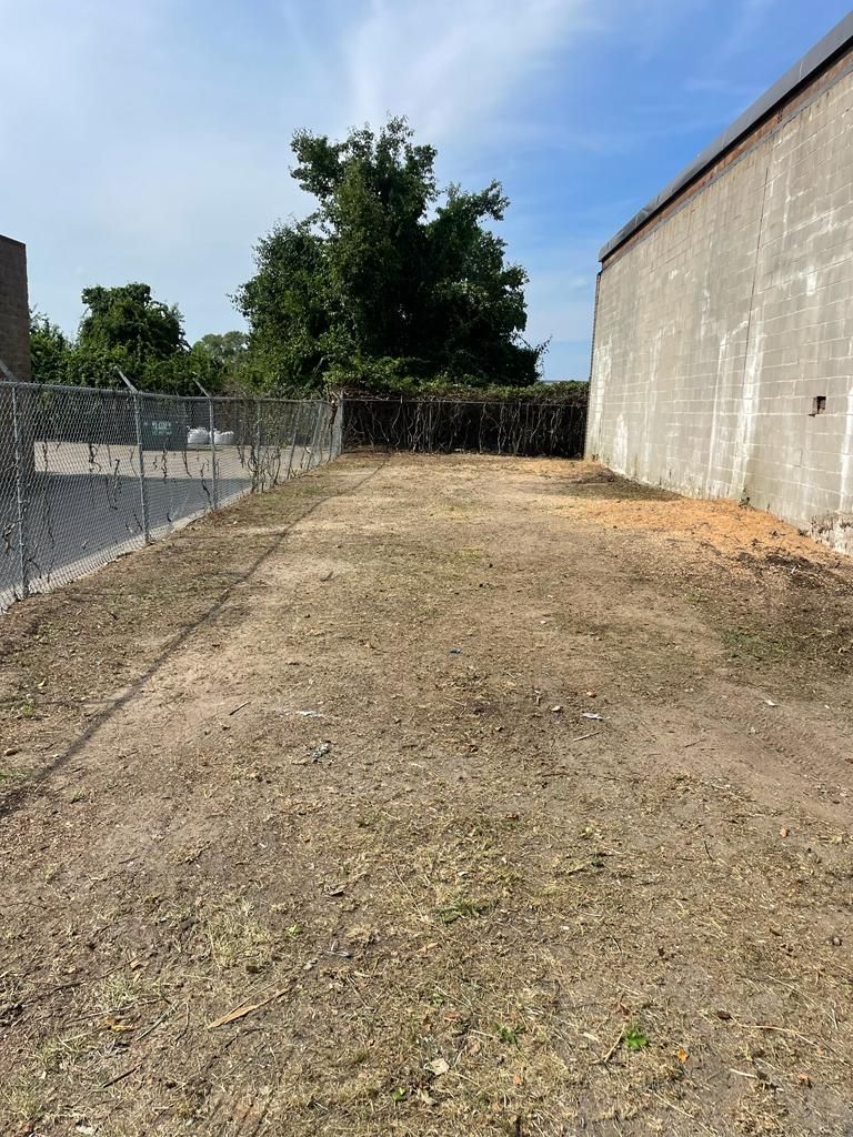Empty gravel lot beside a beige building, fenced on the left, with trees and blue sky in back.