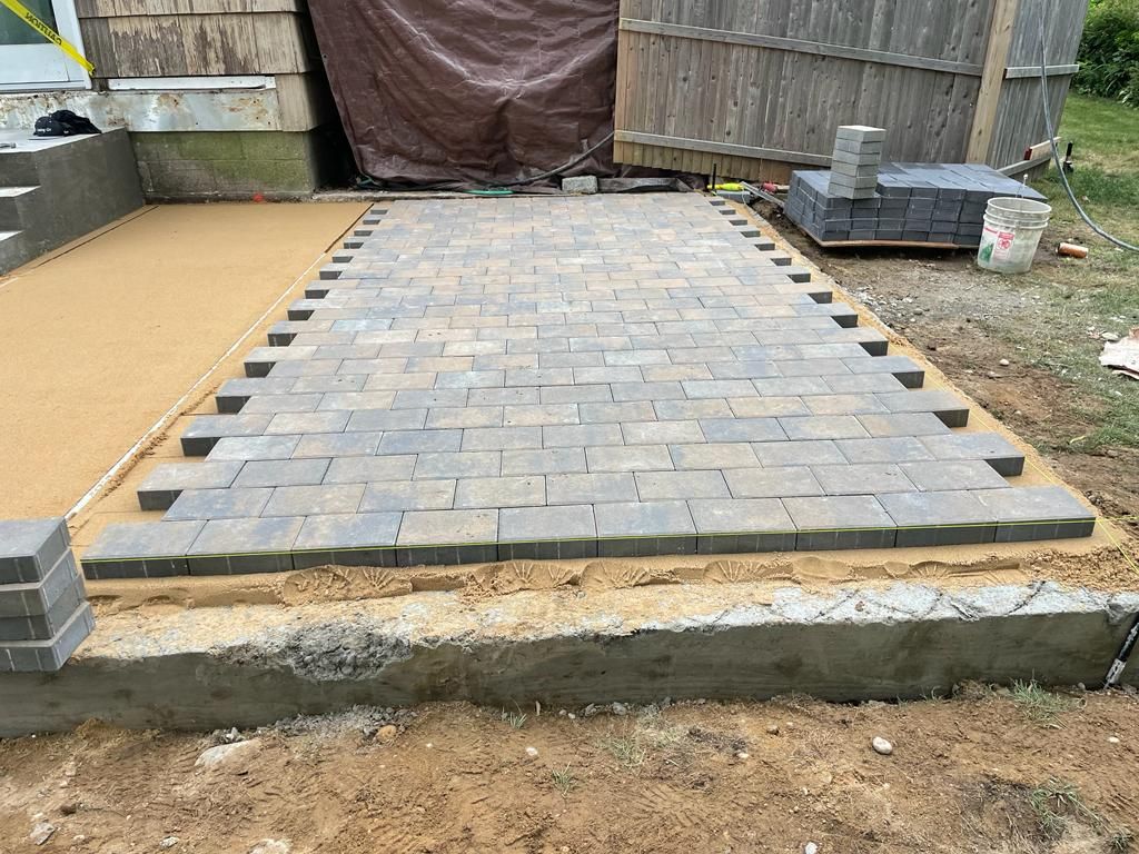 Partially paved backyard patio with gray pavers, concrete edging, and stacked blocks at the sides