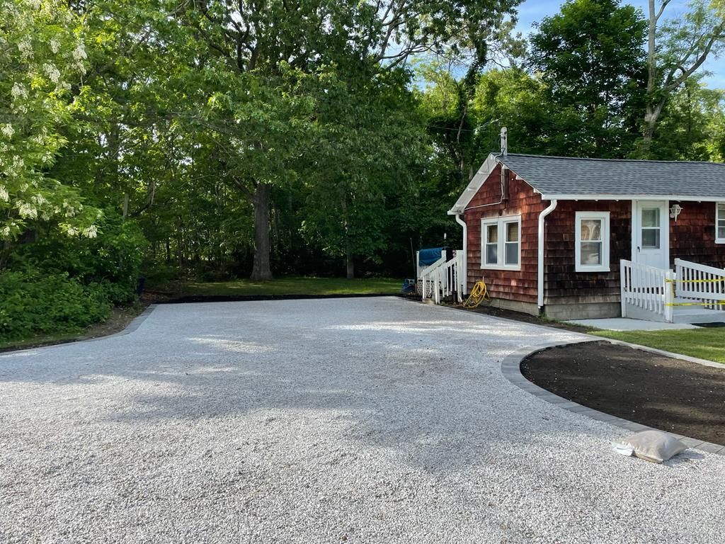 Gravel driveway beside a small red cottage, with white trim and trees in the background.