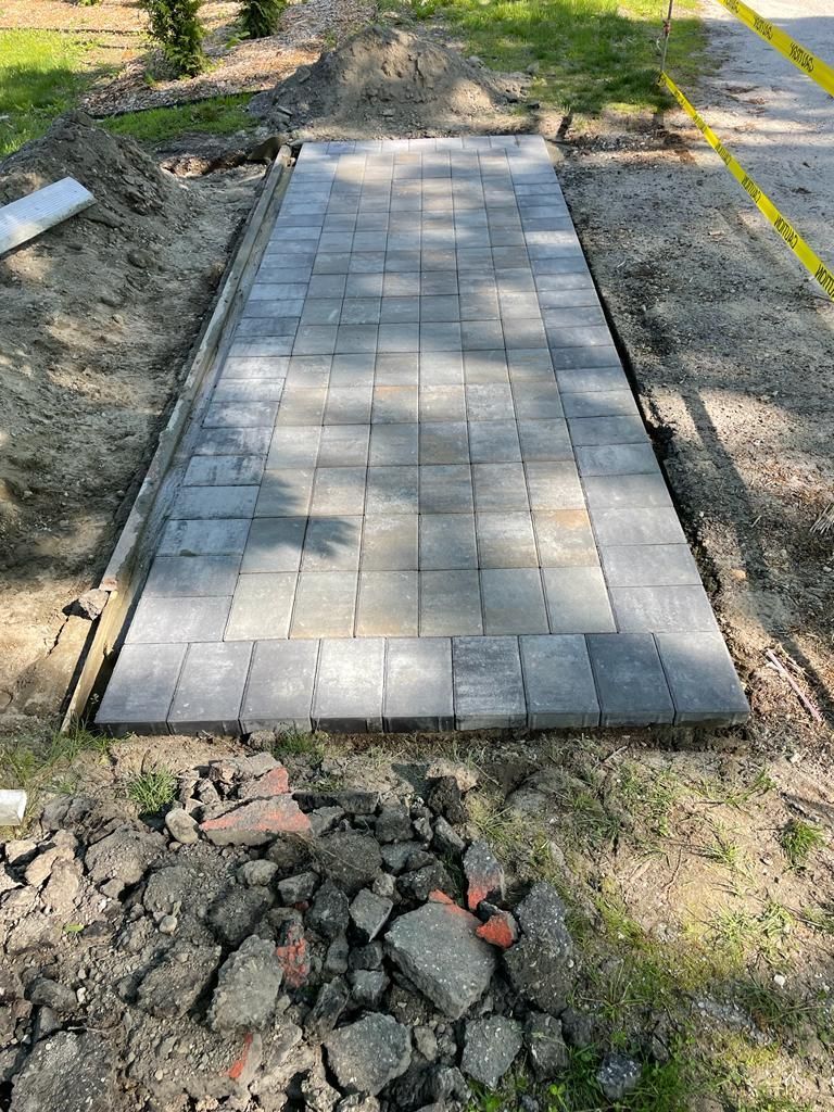 New gray paver walkway section under construction, bordered by dirt and gravel.