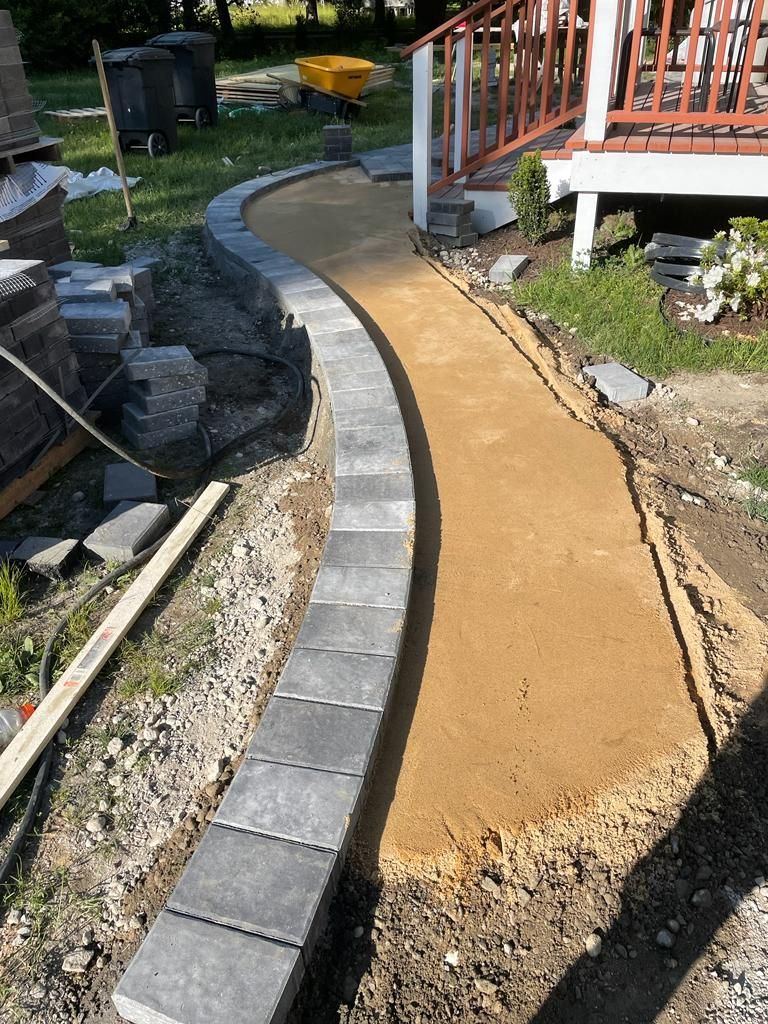 Curved walkway under construction beside a porch, with stone edging and a gravel path in a yard