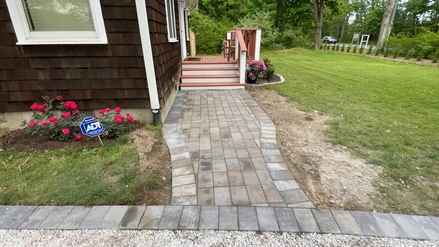 Stone walkway leading to a brown house porch, with flower beds and a grassy yard on the side