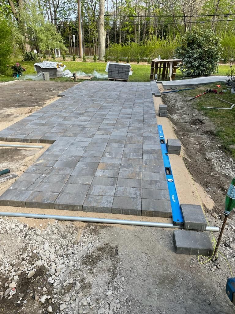 Stone paver patio under construction in a backyard, with edging and leveling tools visible.