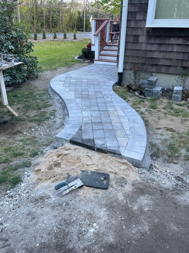 Curved gray paver walkway under construction beside a house, with a dirt yard and stairs ahead