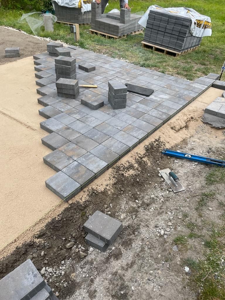 Partially laid gray paver patio with loose blocks and tools in a yard