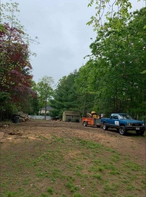 Dirt lot with a blue truck, orange tractor, and trees under an overcast sky