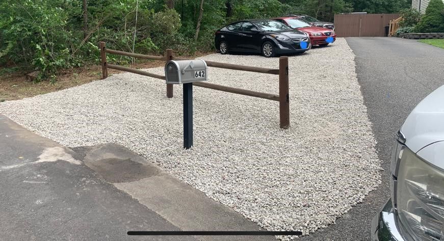 Gravel parking area with a mailbox, wooden barrier, and parked cars beside a wooded road.