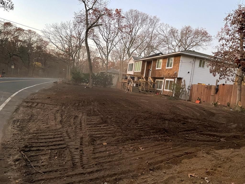 Dirt lot beside a curved road, with a two-story brick house and bare trees in late afternoon light