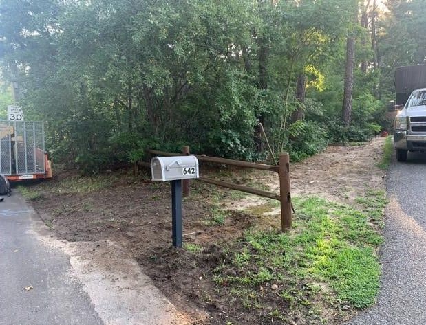 Mailbox beside a driveway with a wooden fence, trees, and a parked truck in the background.