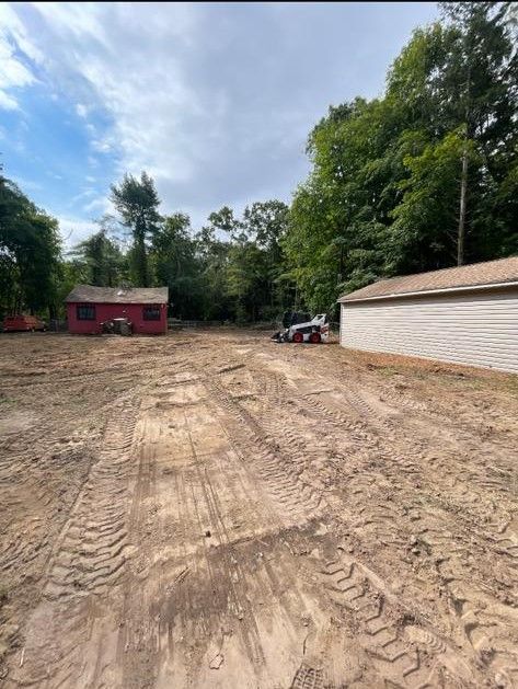 Cleared dirt lot beside a white garage and red shed under a partly cloudy sky