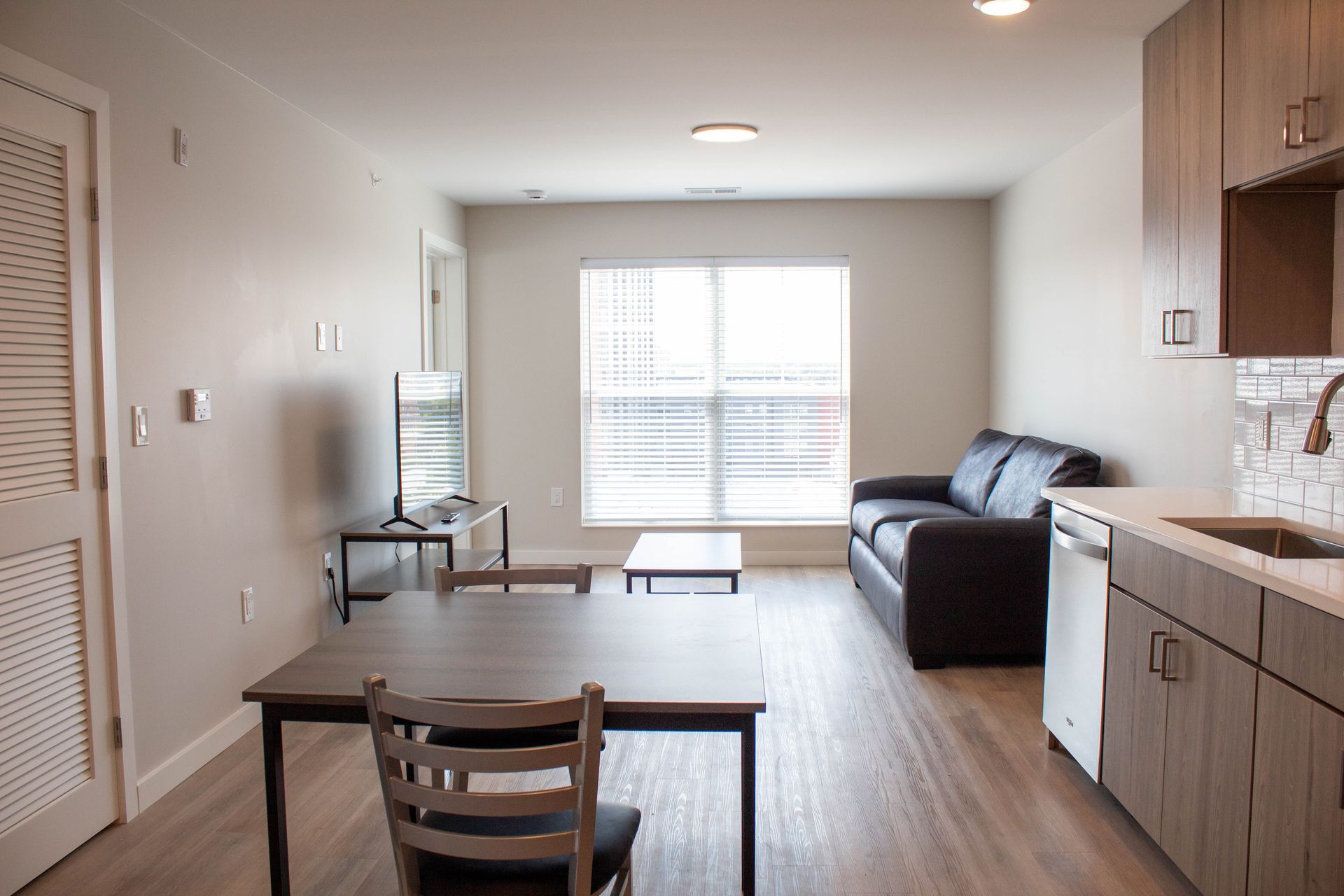 Interior of a modern apartment; living area, kitchen, dining table, sofa, TV, and window.