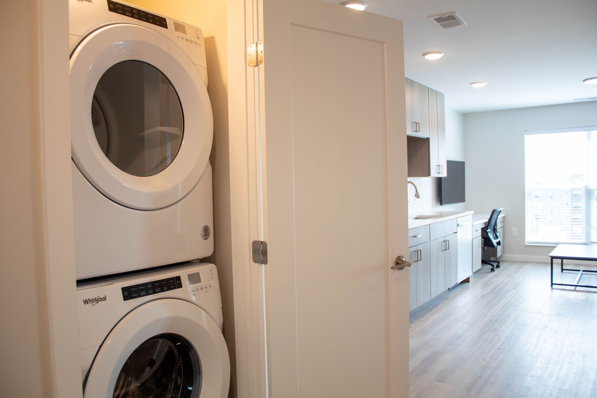 Stacked white washer and dryer in a small laundry area. White door leads to a modern living space.