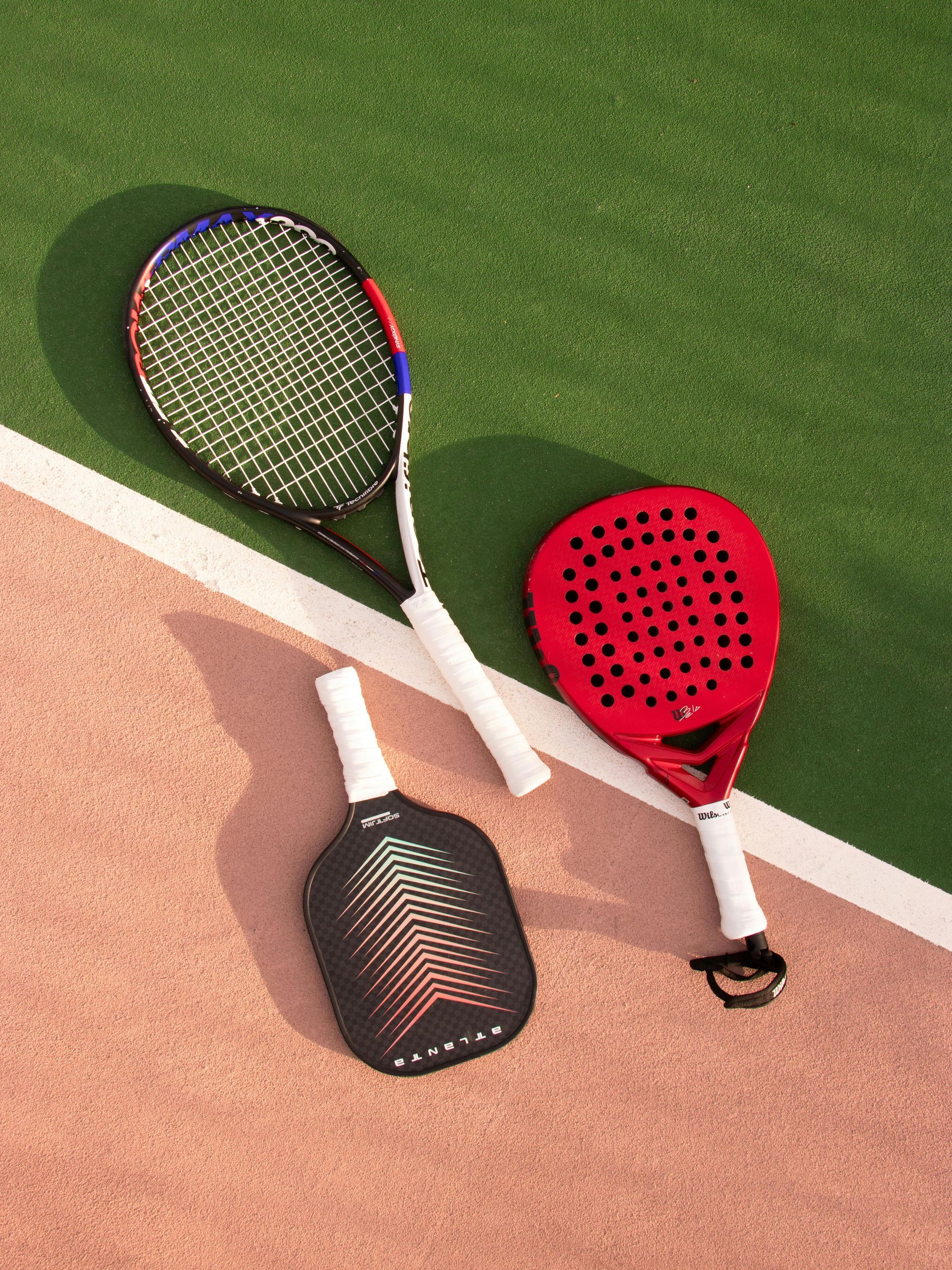 Three padel rackets on a court. One is red, one black, and one has blue accents, all with white handles.