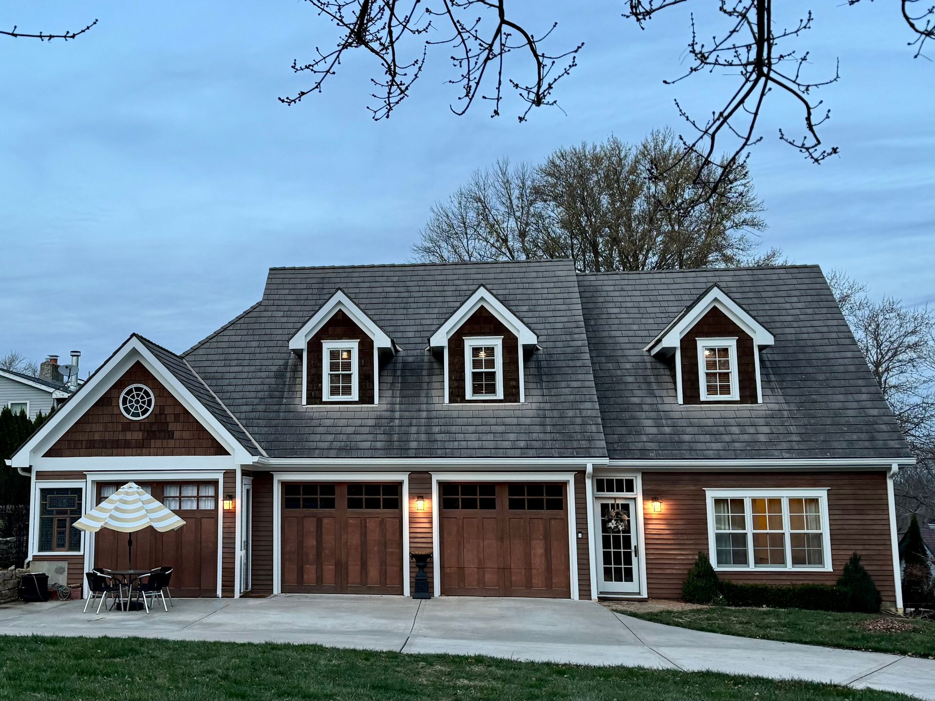 Brick suburban house with gray roof, three dormer windows, and lit front porch at dusk